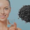 Alt : Femme souriante avec un verre d’eau et un charbon actif en gros plan, mettant en avant des solutions de nettoyage écologique pour un intérieur sain.